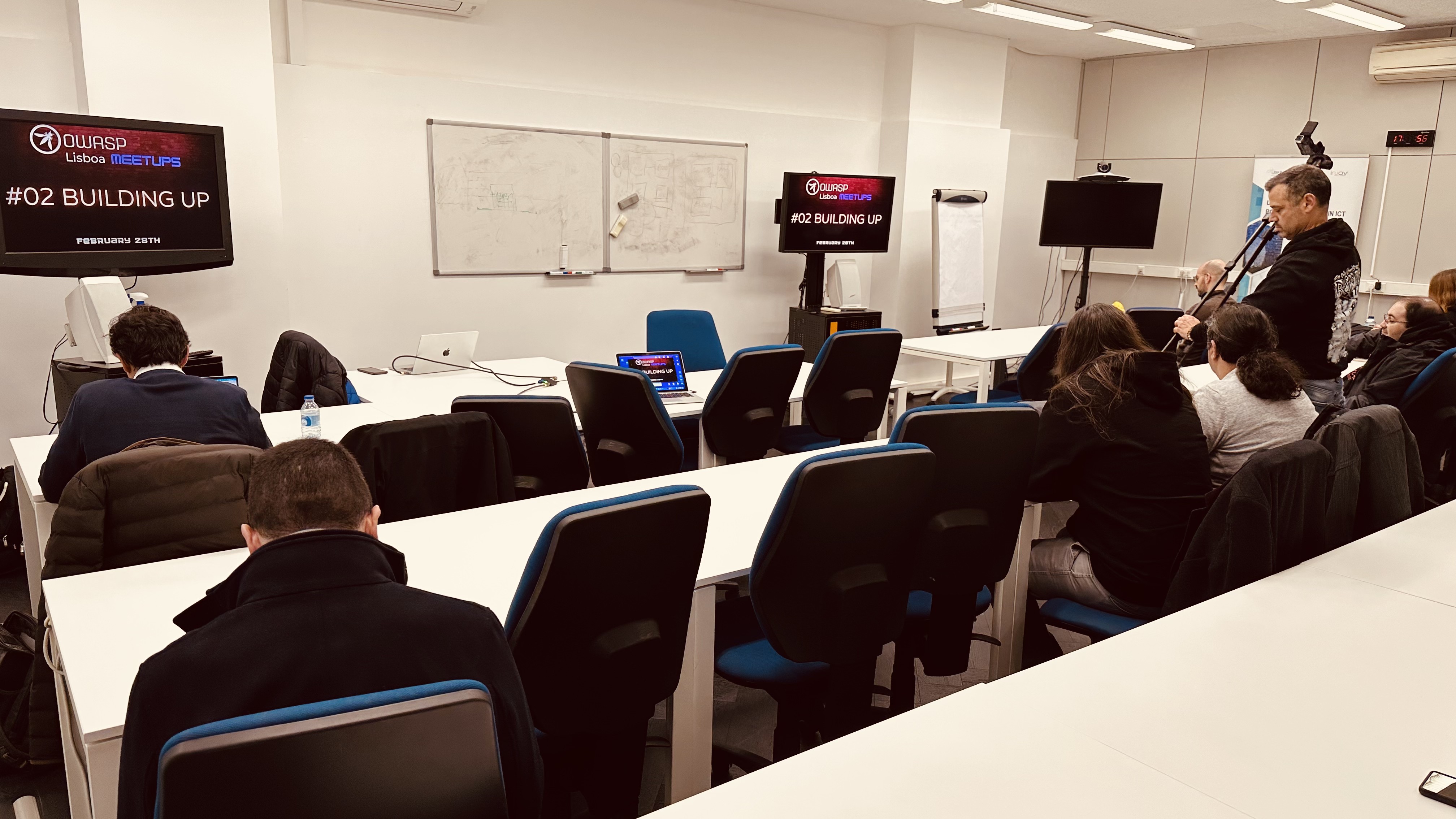 OWASP Lisboa #2 Audience members seated at white tables facing two screens displaying the "OWASP Lisboa Meetups #02 Building Up" title slide, with a videographer recording the session from the side.