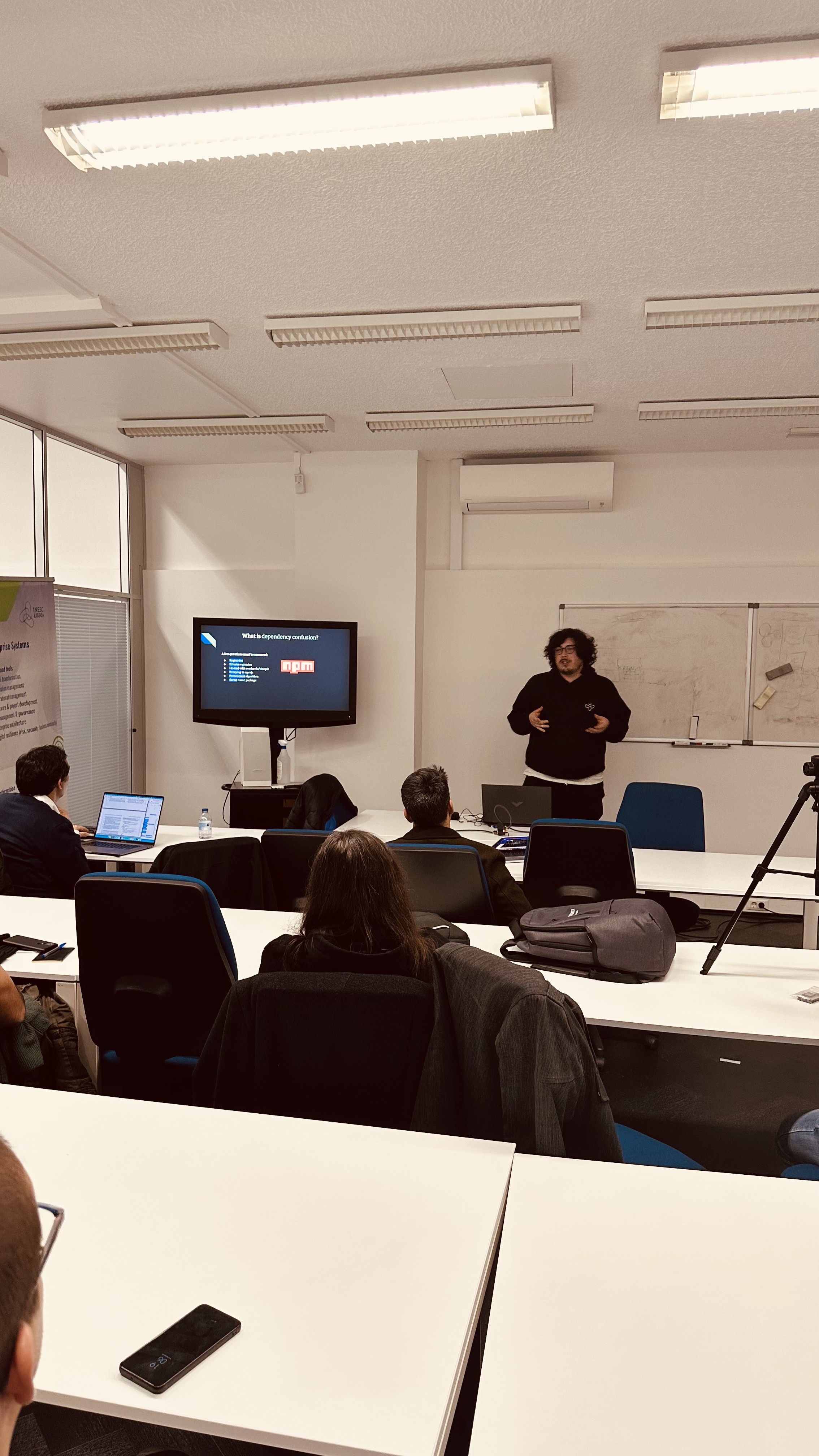 OWASP Lisboa #2 The speaker in the black hoodie presenting a technical slide featuring the npm logo, viewed from behind the seated audience.