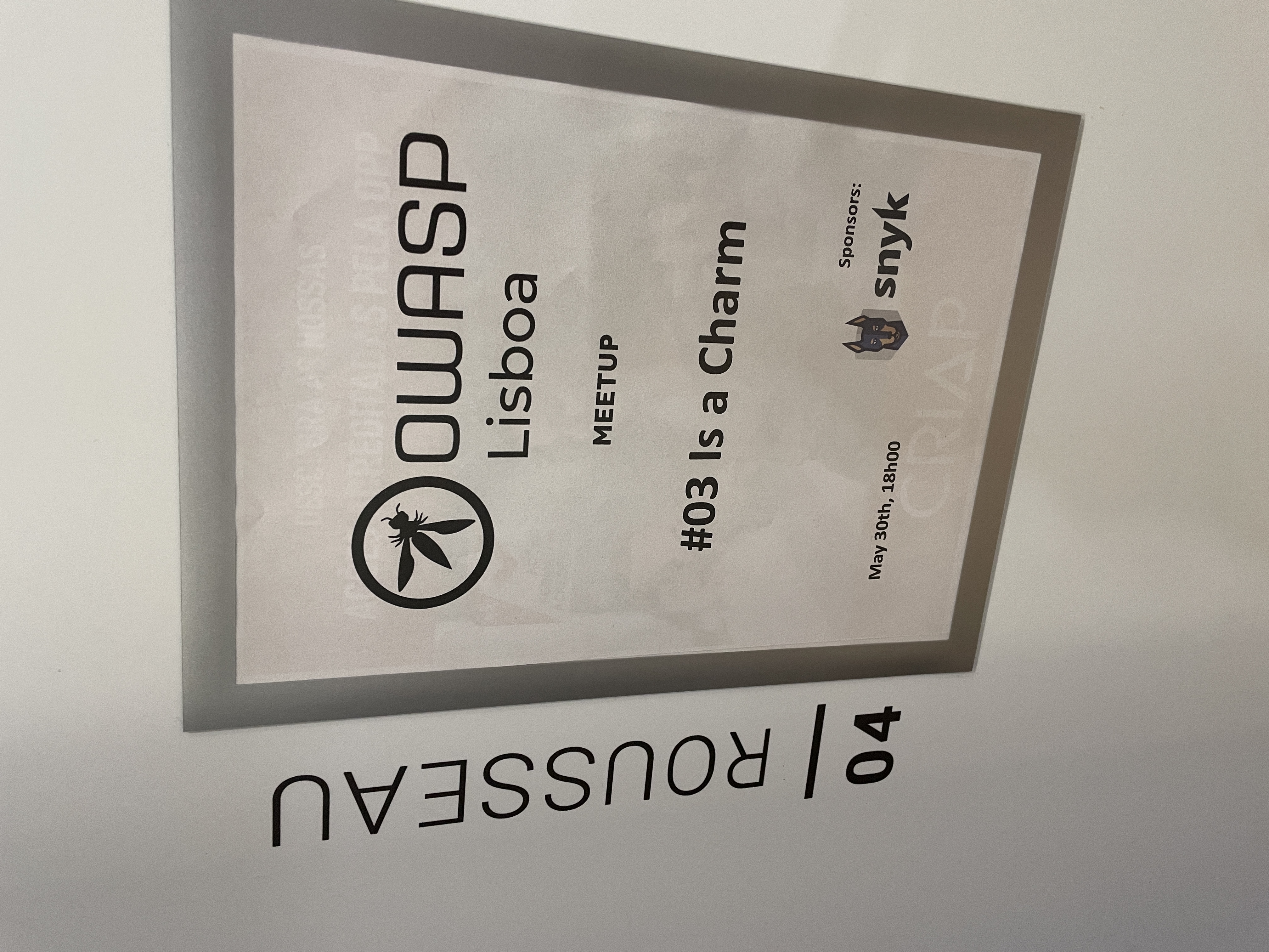 OWASP Lisboa #3 A printed event sign posted on a wall next to room "04 Rousseau," displaying the OWASP Lisboa logo, the event title "#03 Is a Charm," and the sponsor logo for Snyk.