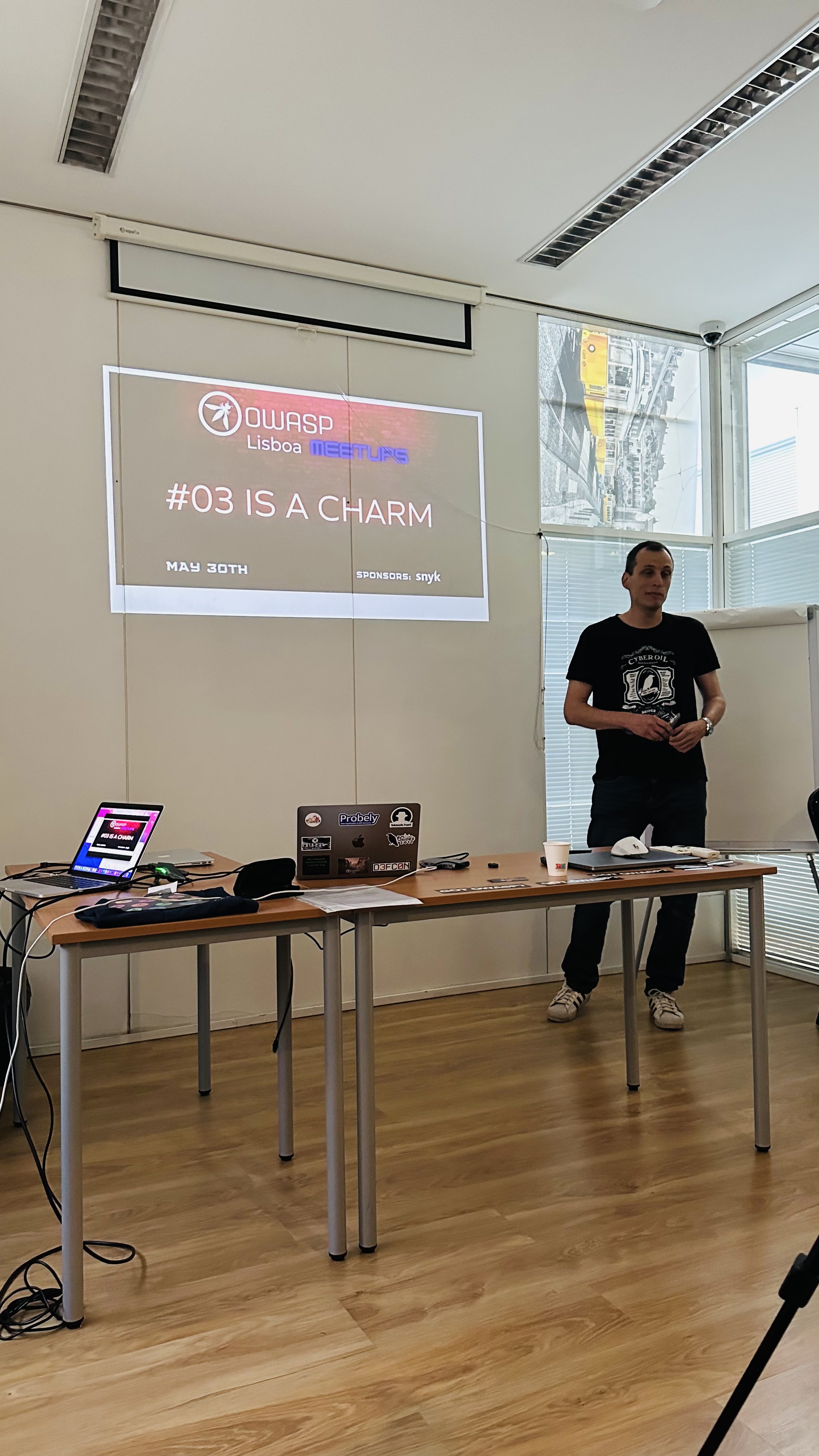 OWASP Lisboa #3 A speaker standing next to a projection screen displaying the title slide "OWASP Lisboa Meetups #03 IS A CHARM" with the date May 30th.