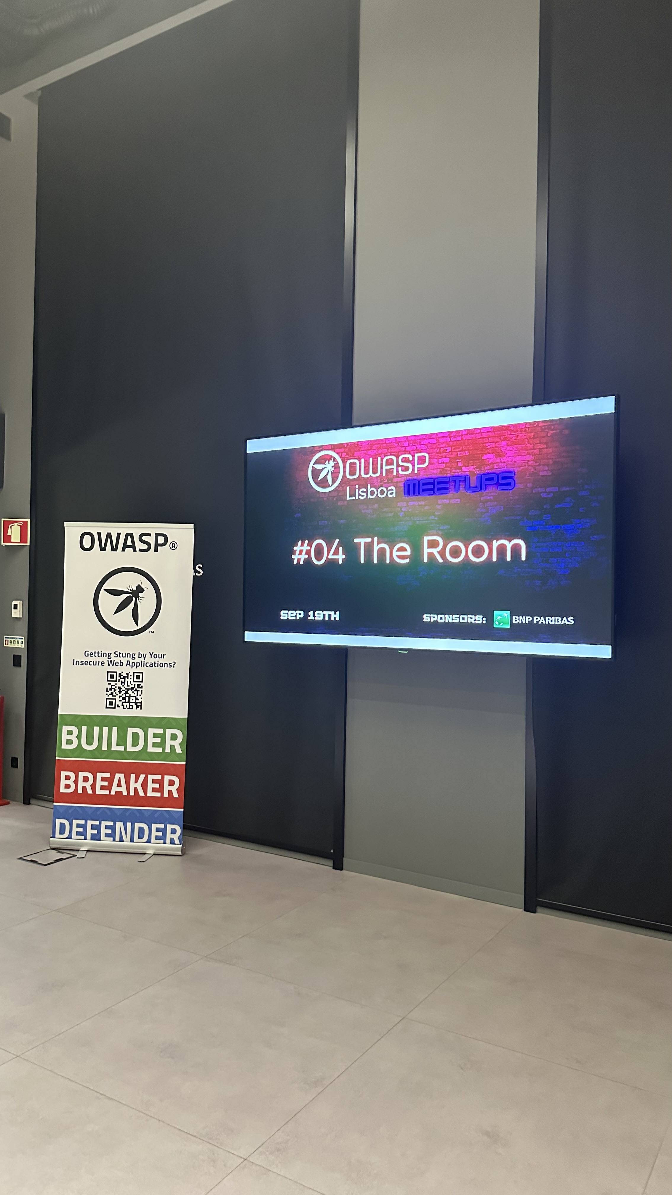 OWASP Lisboa #4 A vertical OWASP banner standing next to a large wall-mounted screen displaying the event title "#04 The Room" and the sponsor logo.