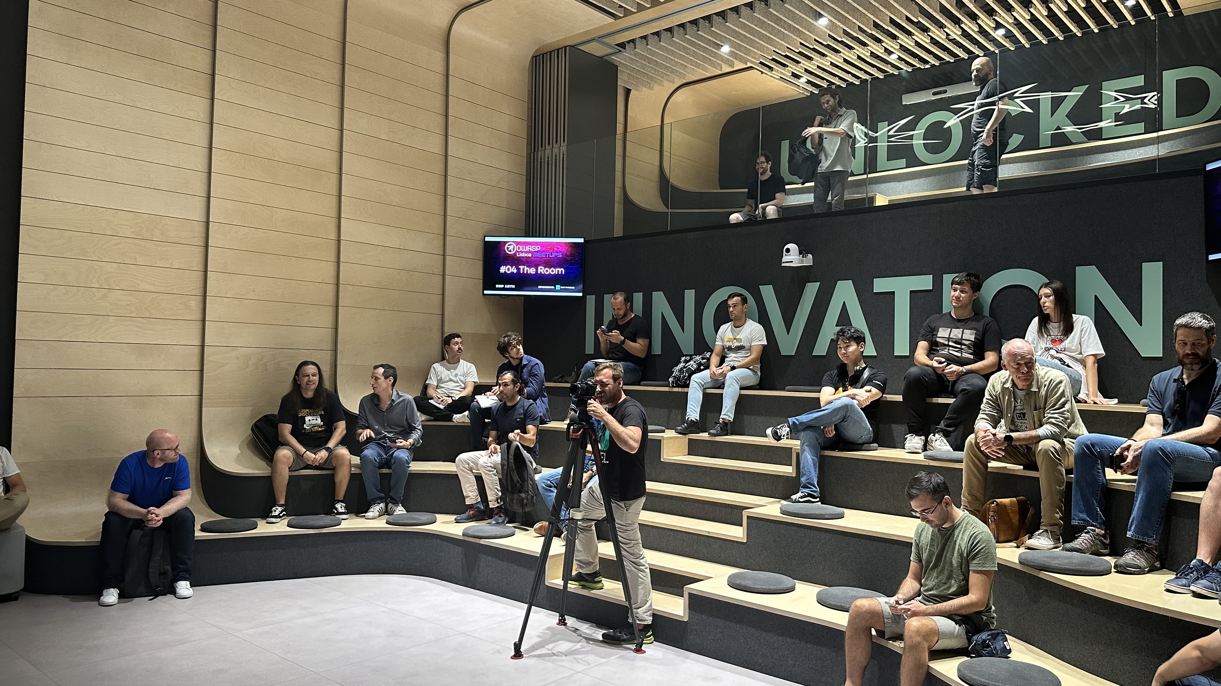 OWASP Lisboa #4 Attendees seated on tiered wooden benches in a modern venue with the word "INNOVATION" written in large letters on the wall behind them.