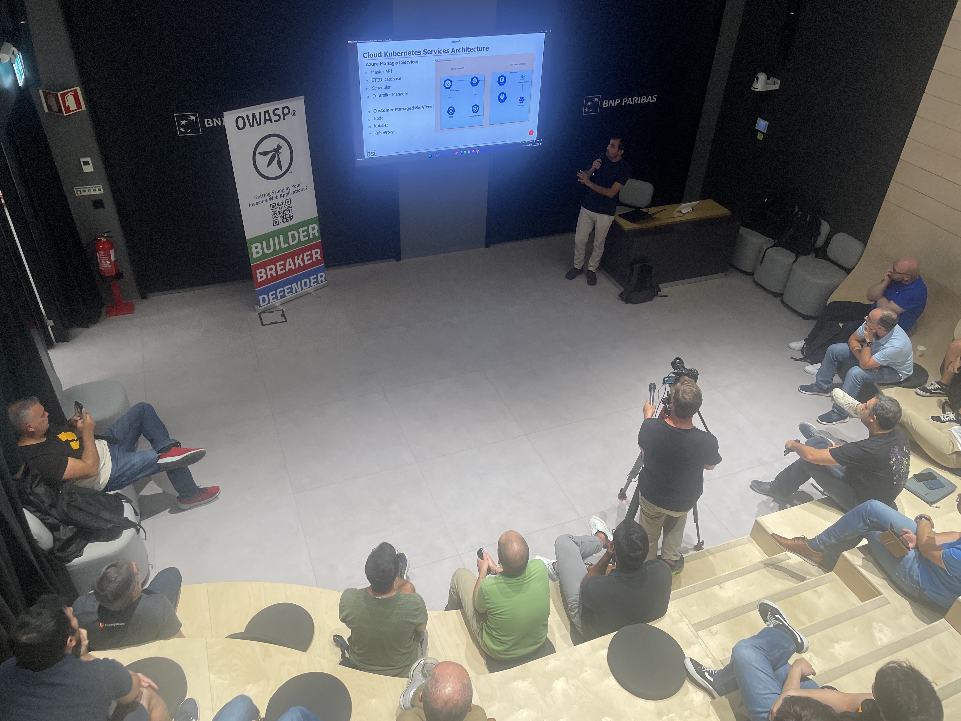 OWASP Lisboa #4 An overhead view of the audience seated on tiered benches watching a presentation on "Cloud Kubernetes Services Architecture," with a videographer recording the talk.