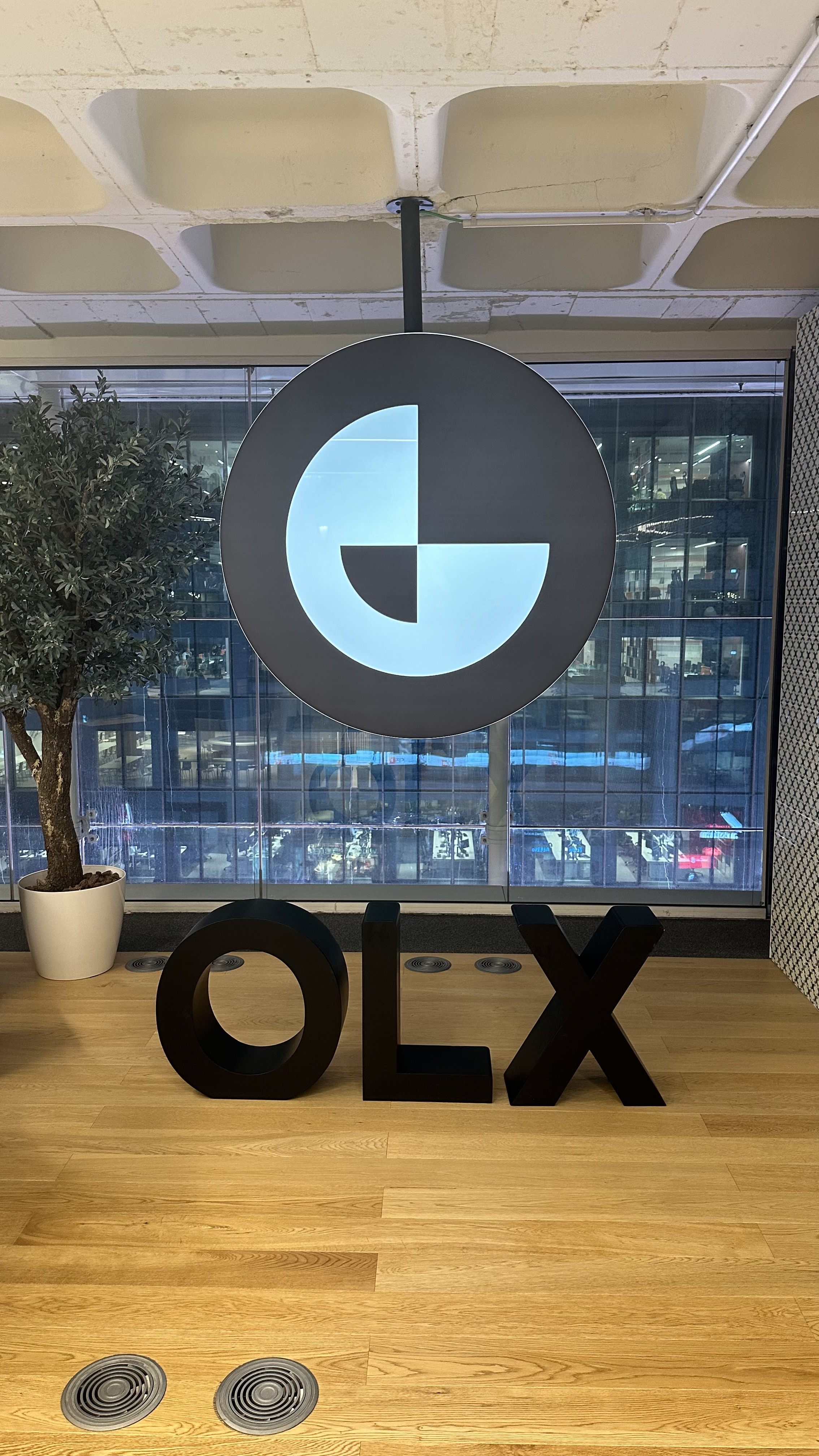 OWASP Lisboa #5 Large black letters spelling "OLX" on the wooden floor and a hanging circular logo in a modern office lobby with glass walls overlooking the city.