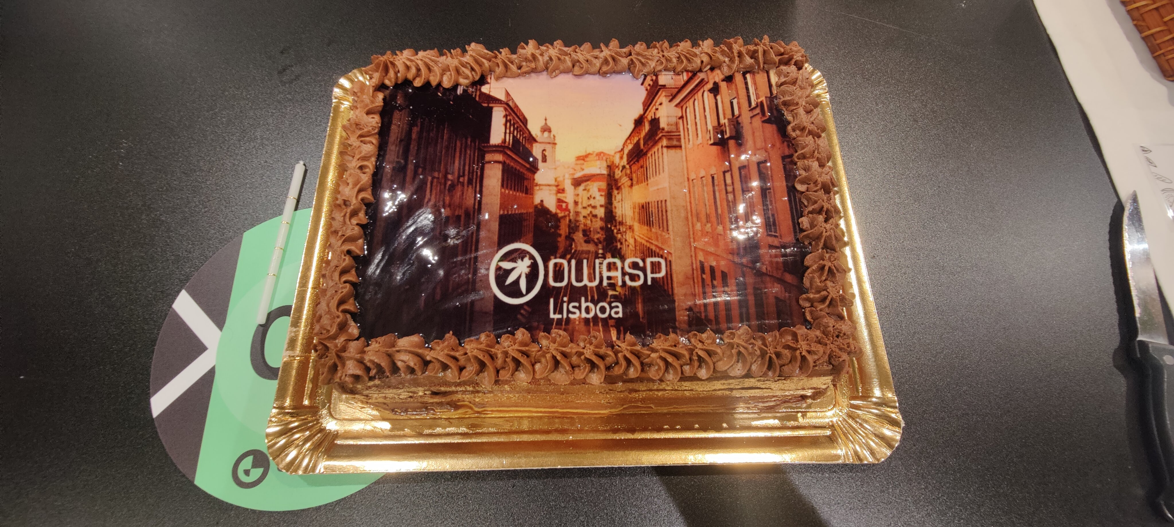 OWASP Lisboa #5 A rectangular celebratory cake topped with an edible photo of a Lisbon street and the OWASP Lisboa logo, surrounded by chocolate piping.