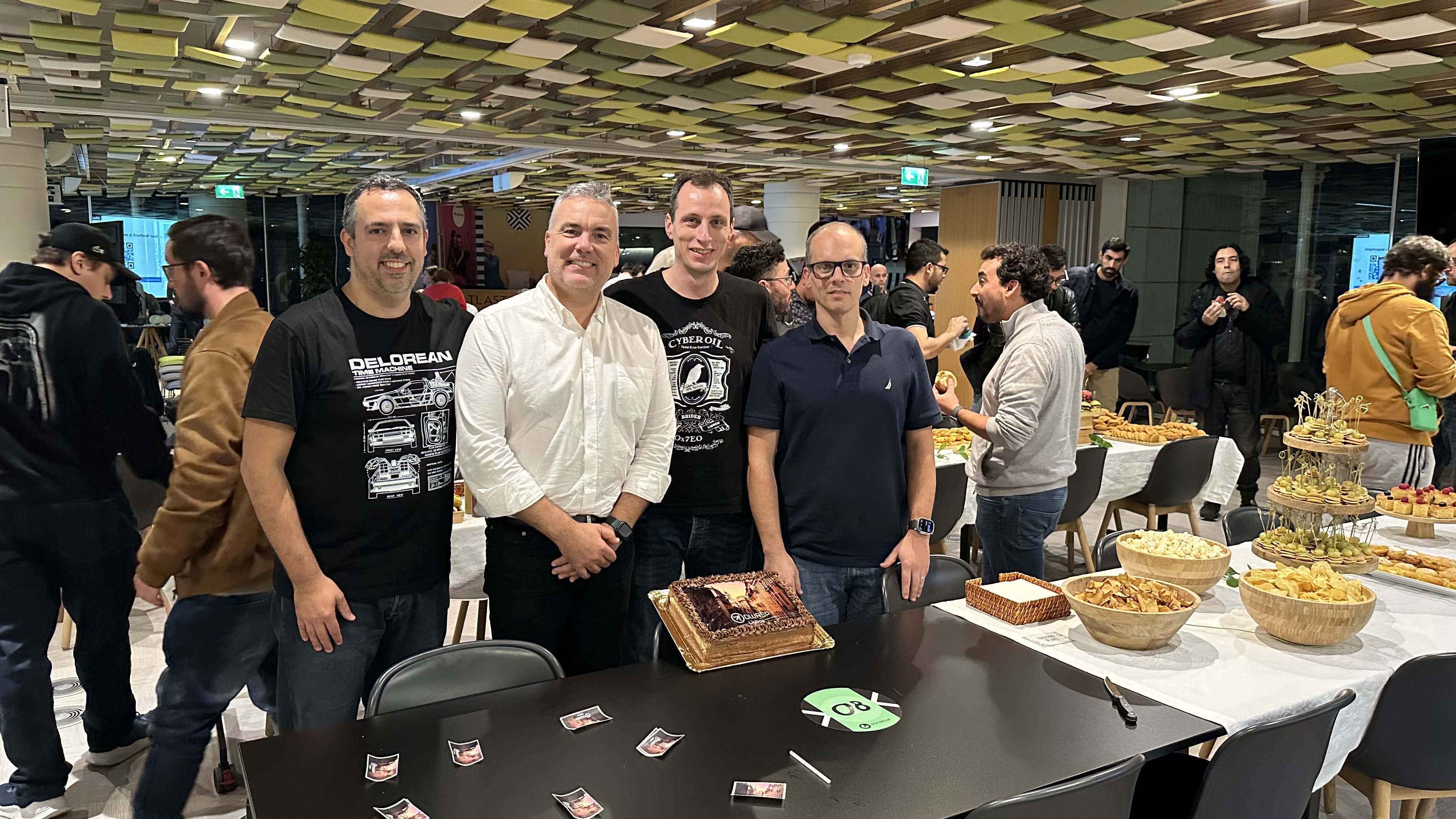 OWASP Lisboa #5 Four chapter leaders posing for a photo behind a table with the OWASP cake, with event attendees and catering tables visible in the background.