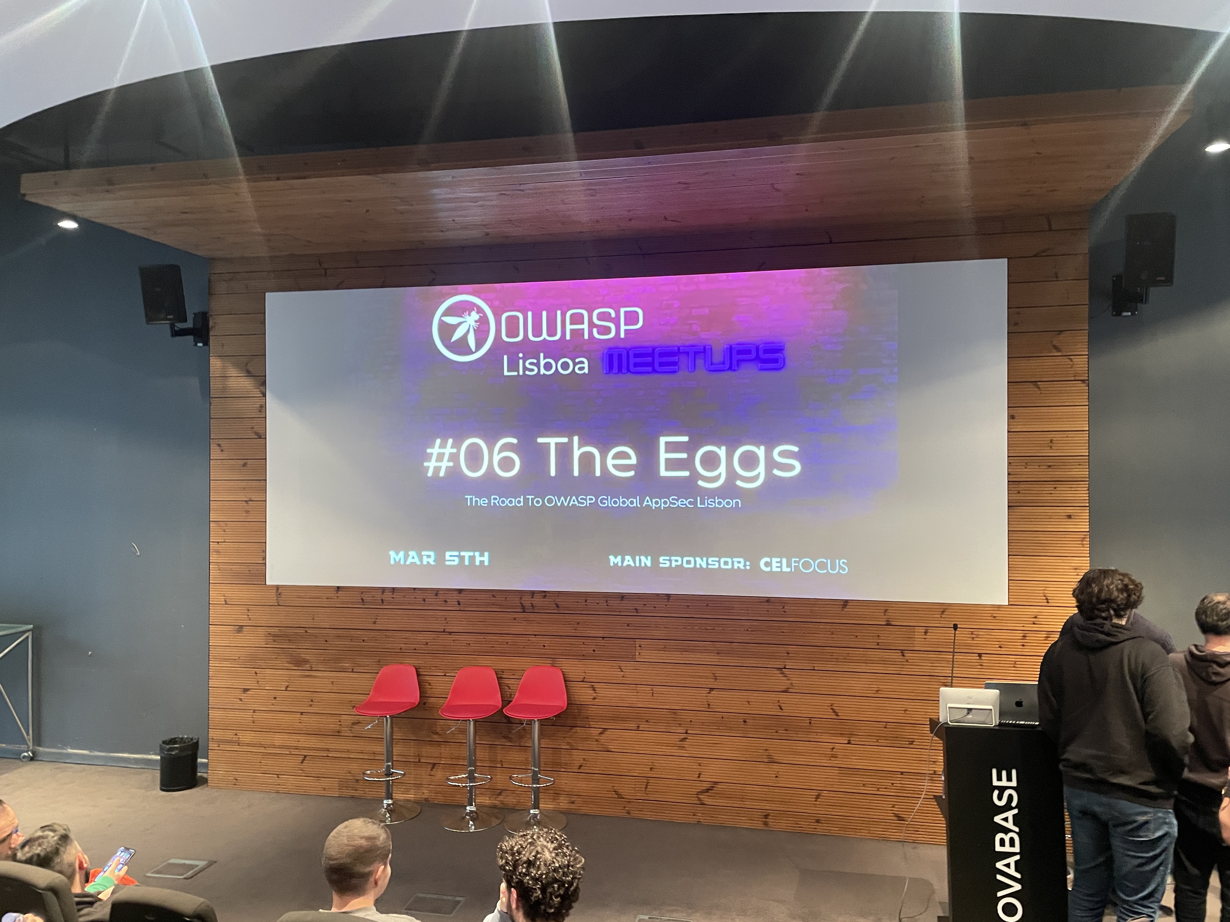 OWASP Lisboa #6 The main stage screen displaying the title slide "OWASP Lisboa Meetups #06 The Eggs" with date and sponsor details, positioned above three red stools.