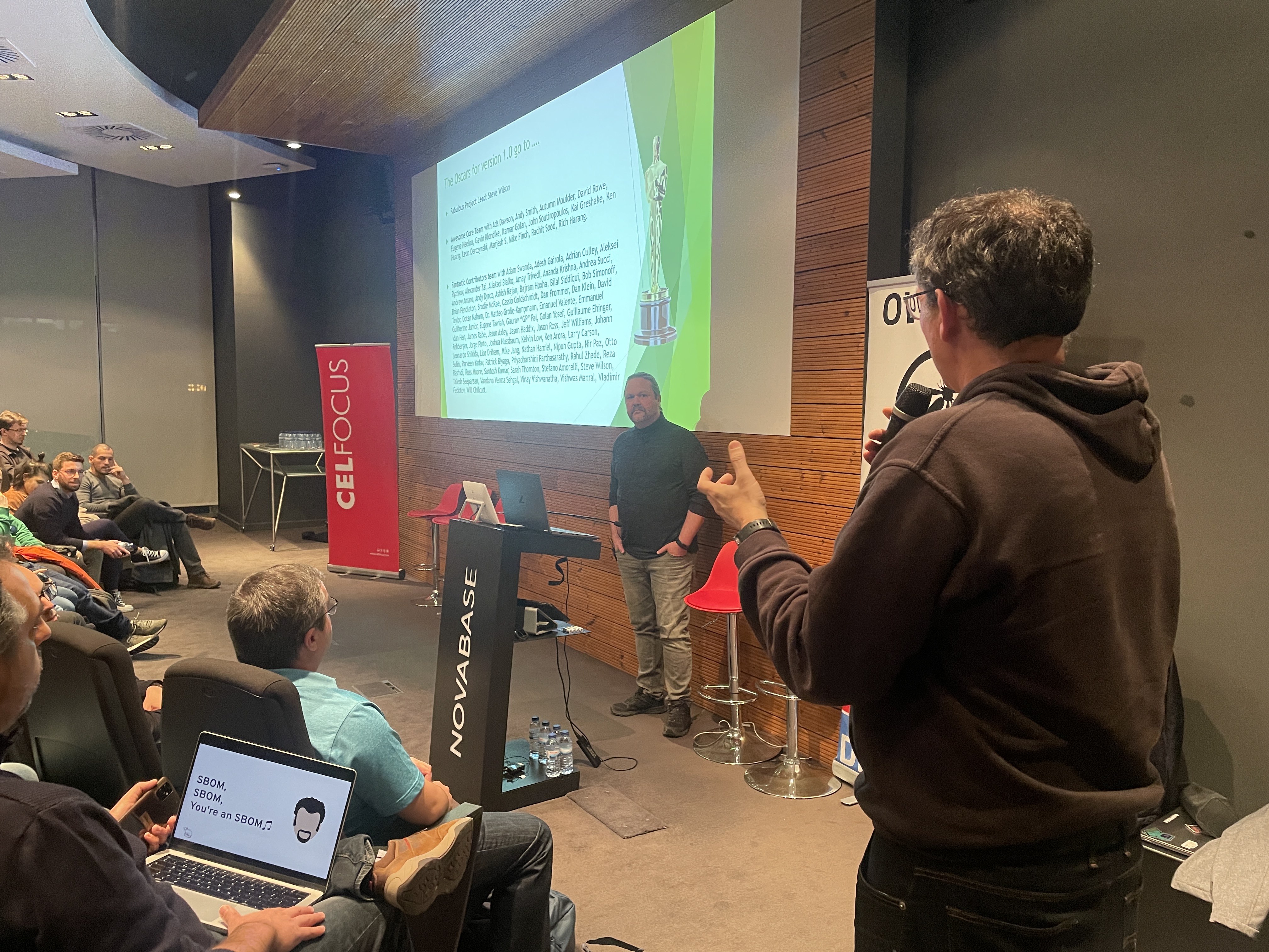 OWASP Lisboa #6 An audience member holding a microphone asks a question to the speaker, with a list of contributors displayed on the main screen.