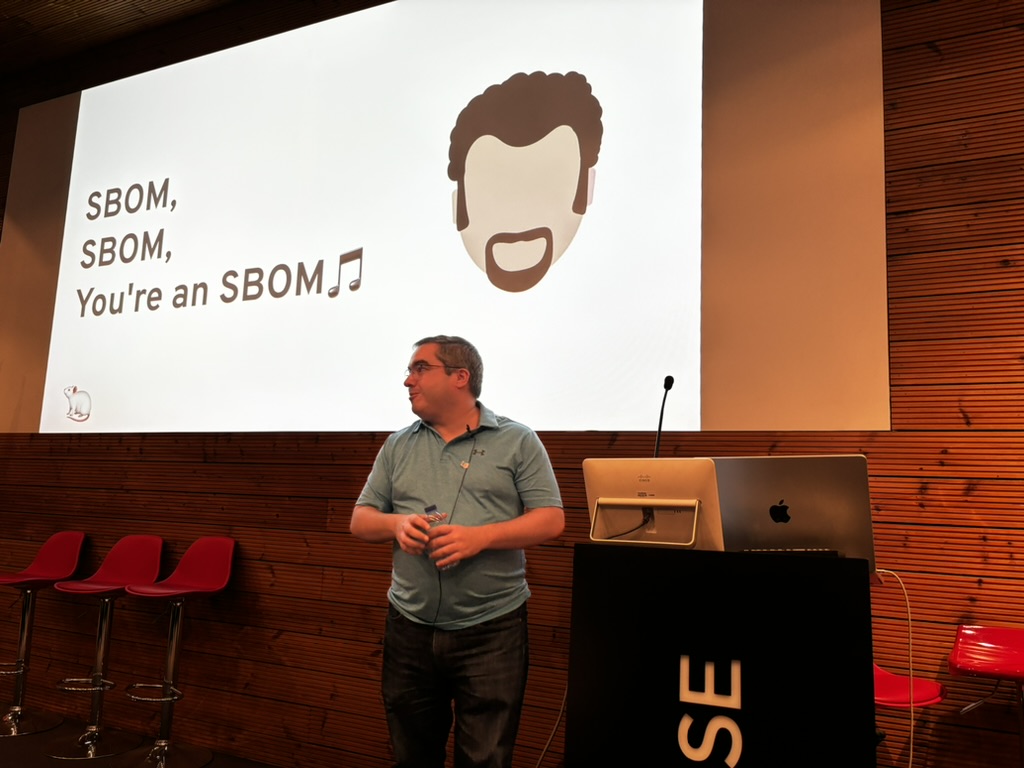 OWASP Lisboa #6 A speaker at the podium smiling in front of a humorous slide reading "SBOM, SBOM, You're an SBOM" with a cartoon illustration.