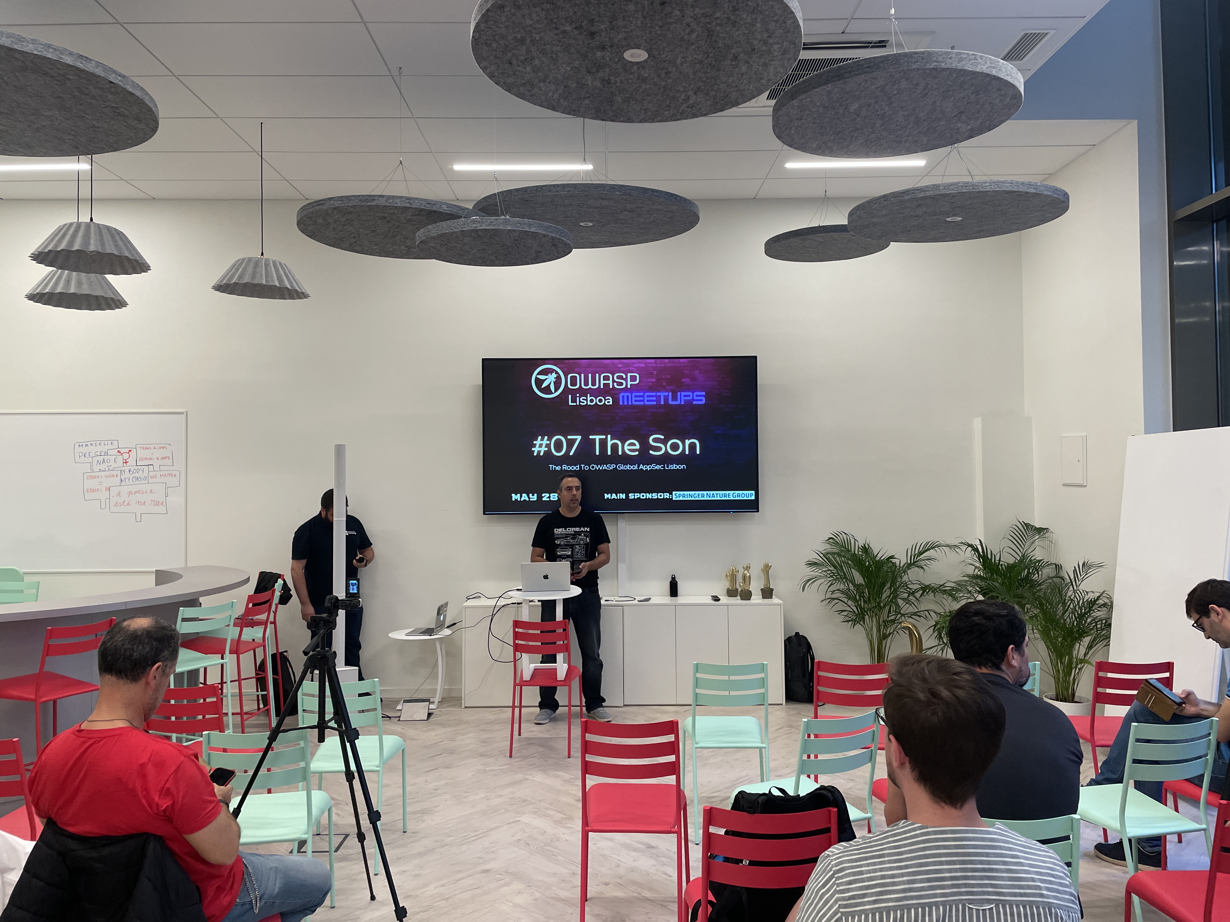 OWASP Lisboa #7 A wide shot of the meetup room with attendees seated on red and mint green chairs, facing a speaker and a screen displaying the "OWASP Lisboa Meetups #07 The Son" title slide.