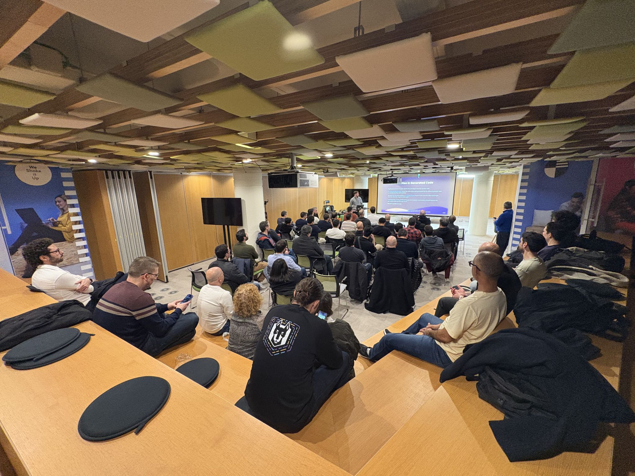 OWASP Lisboa #8 A high-angle view of the audience seated on tiered wooden benches and chairs, listening to a presentation in a room with a colorful acoustic ceiling.