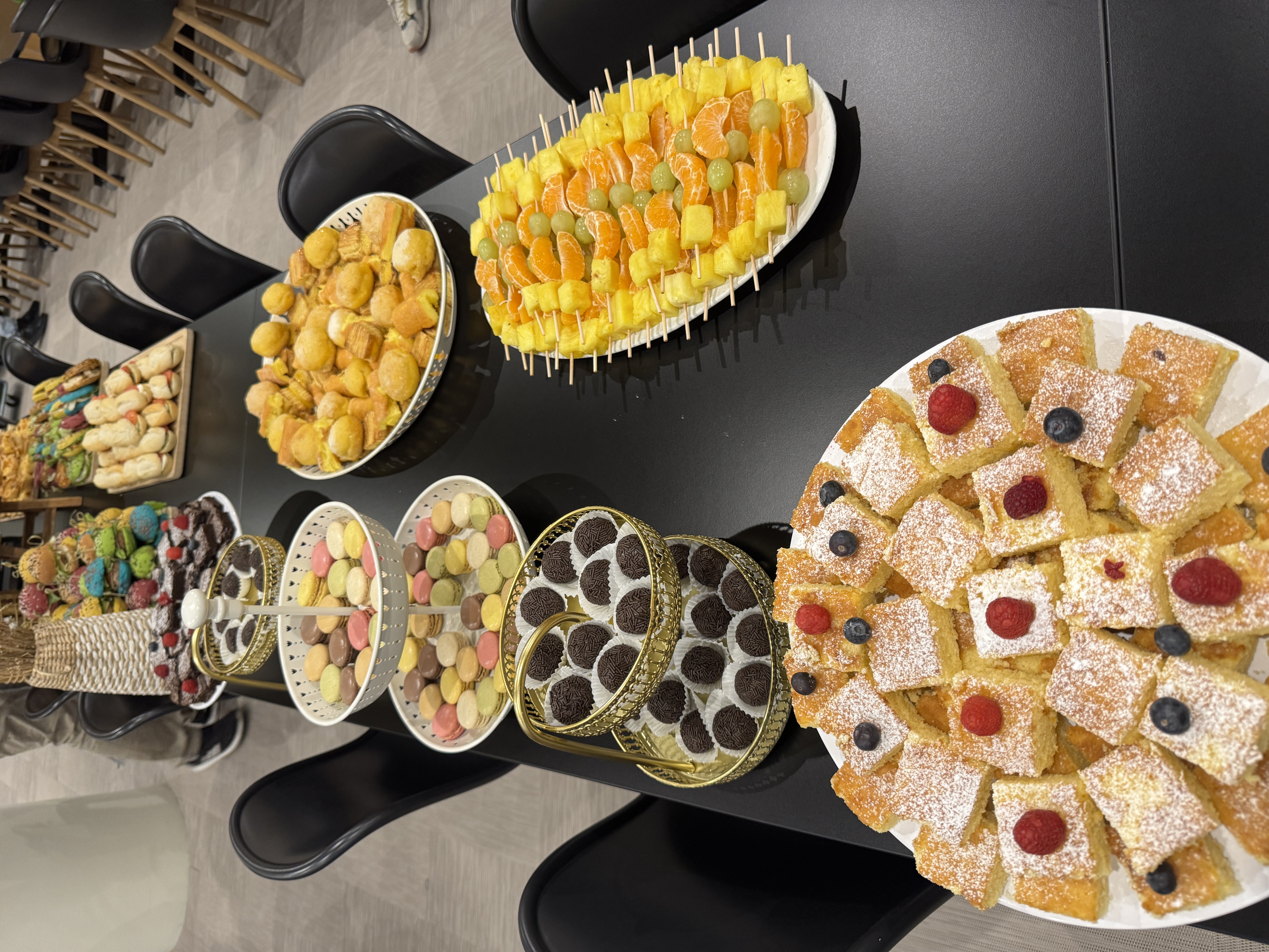 A catering table filled with desserts, including fruit skewers, macarons, chocolate truffles, and sponge cake squares topped with berries.
