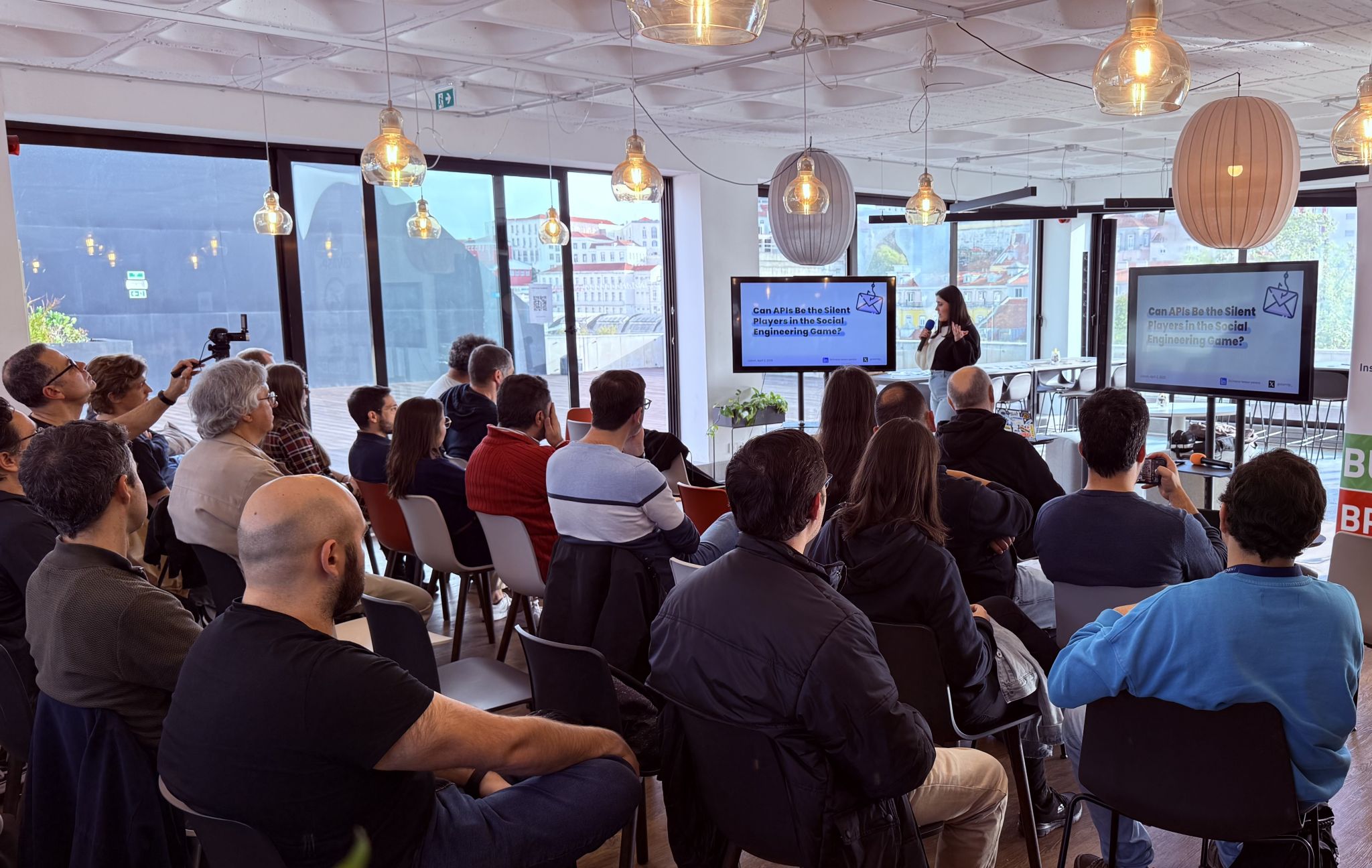 OWASP Lisboa #9 An audience watching a presentation on "Can APIs Be the Silent Players in the Social Engineering Game?" in a room with large windows overlooking the city.