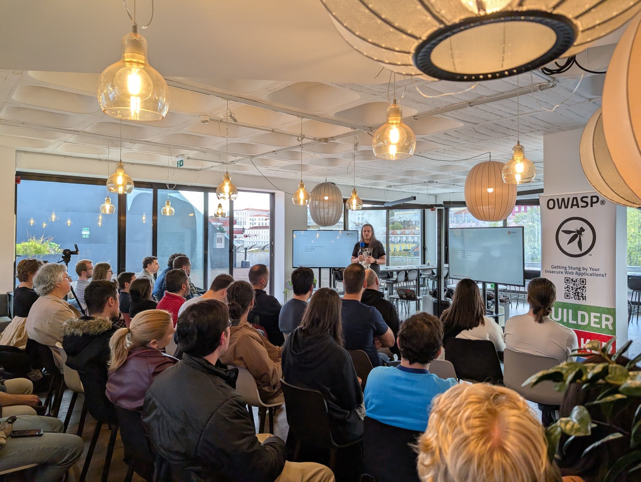 OWASP Lisboa #9 A seated audience listening to a speaker with long hair in a venue featuring modern hanging globe lights and city views.