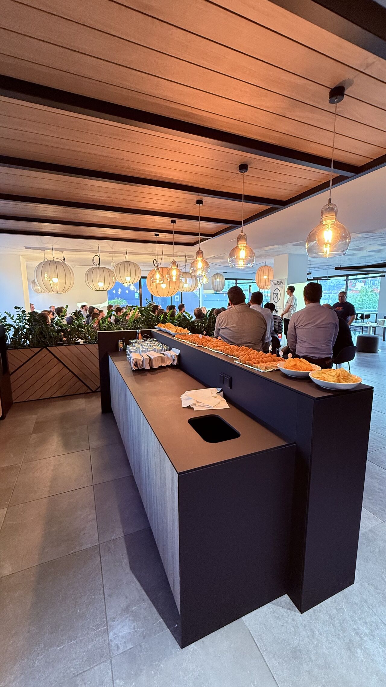 OWASP Lisboa #9 A long catering counter filled with snacks and chips, with attendees networking in the background under warm lighting.