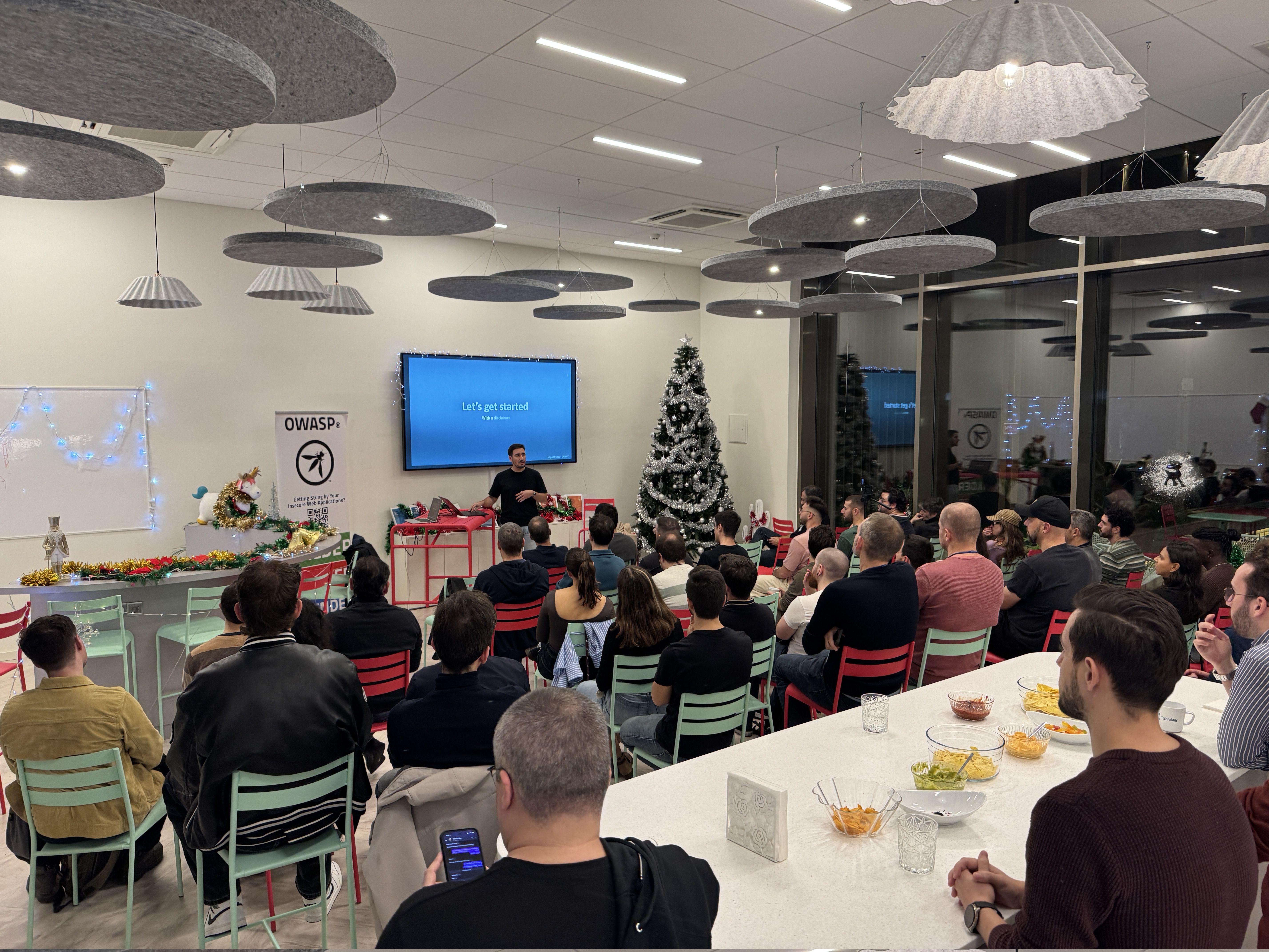 OWASP Lisboa #11 A wide shot of the audience seated in a room decorated for Christmas, listening to a speaker presenting a slide titled "Let's get started."