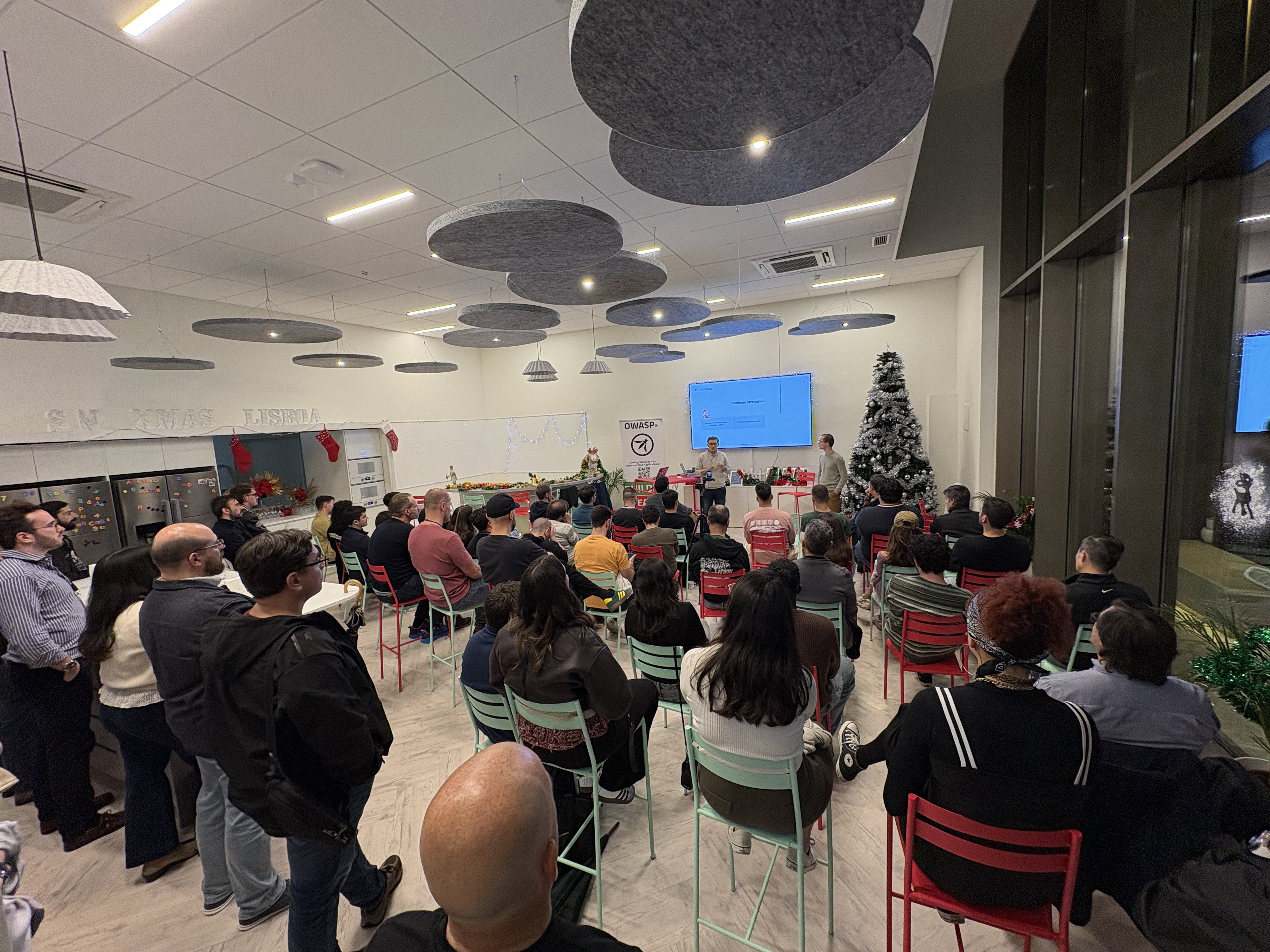 OWASP Lisboa #11 A full view of the meetup room with attendees seated on red and mint green chairs facing the presentation area, which is decorated with festive lights and a tree.