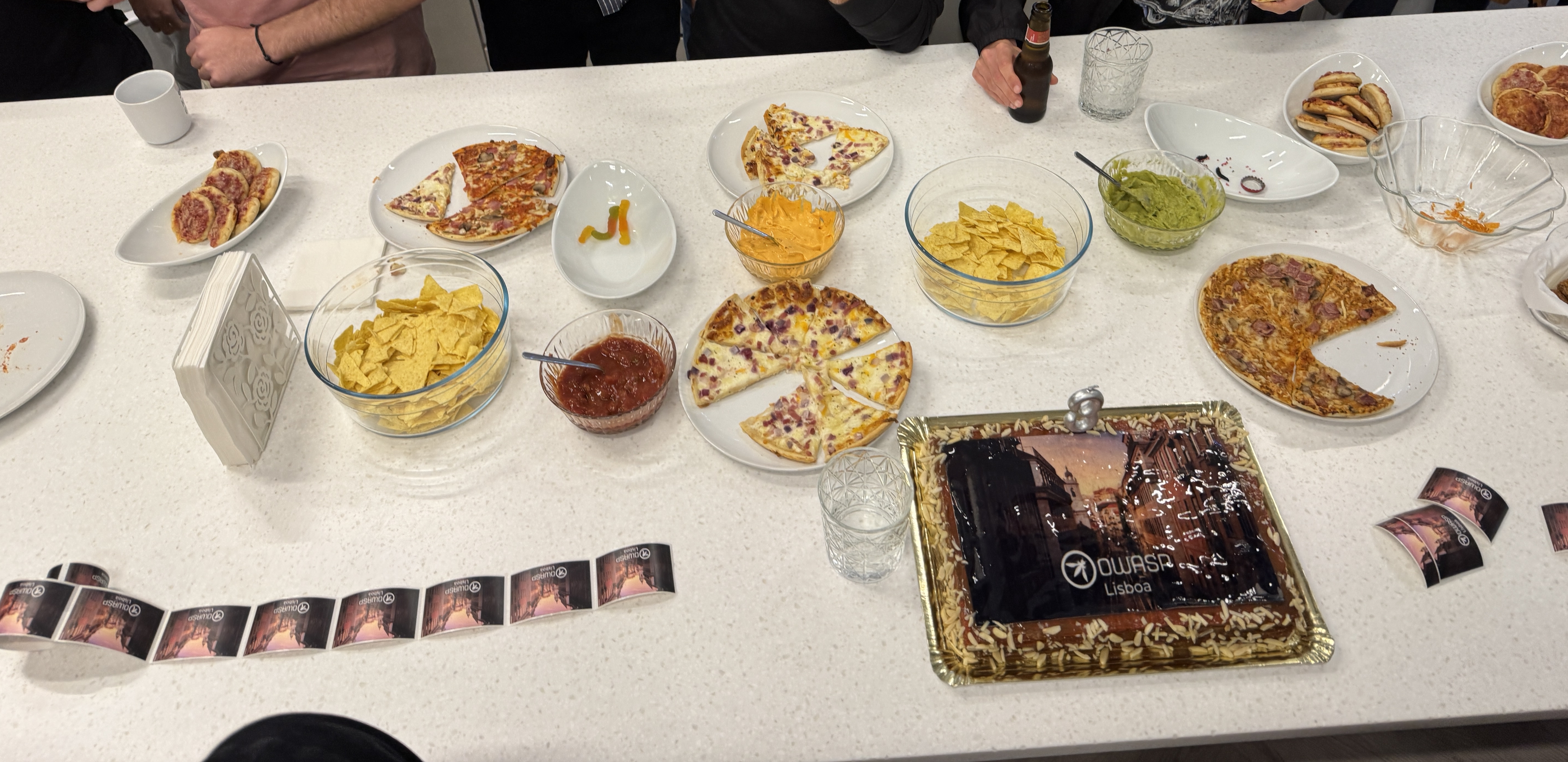 OWASP Lisboa #11 A catering table spread with pizzas, nachos, dips, and a celebratory cake topped with an edible photo of the OWASP Lisboa logo and a "3" candle.