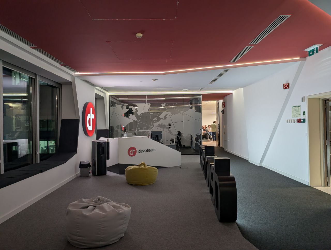 OWASP Lisboa #12 Modern reception area at Devoteam Cybertrust office featuring a white branded desk, a world map graphic on glass walls, and comfortable grey and yellow bean bags.